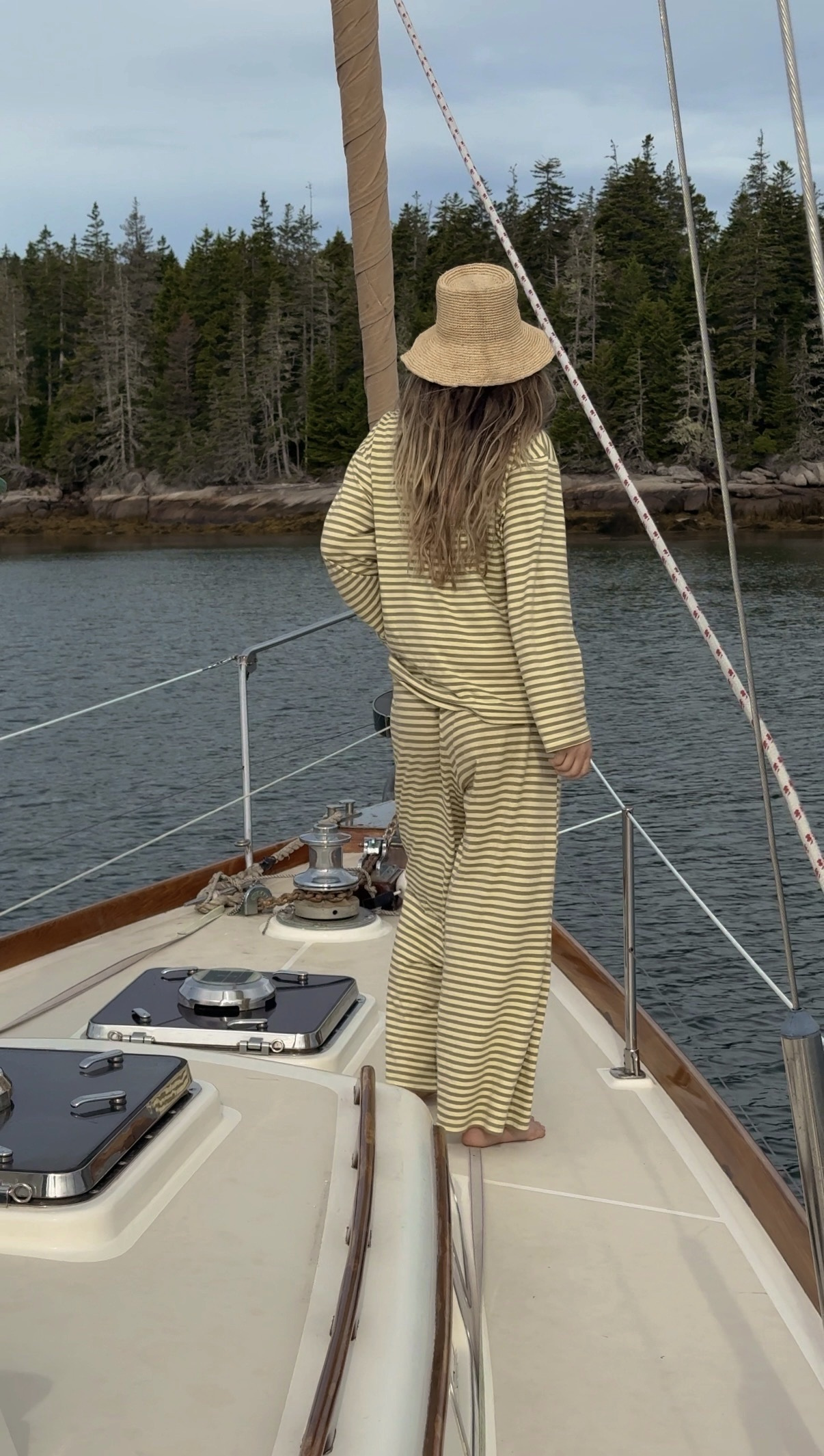 Person in a straw hat and striped outfit aboard a sailboat beside a wooded shoreline